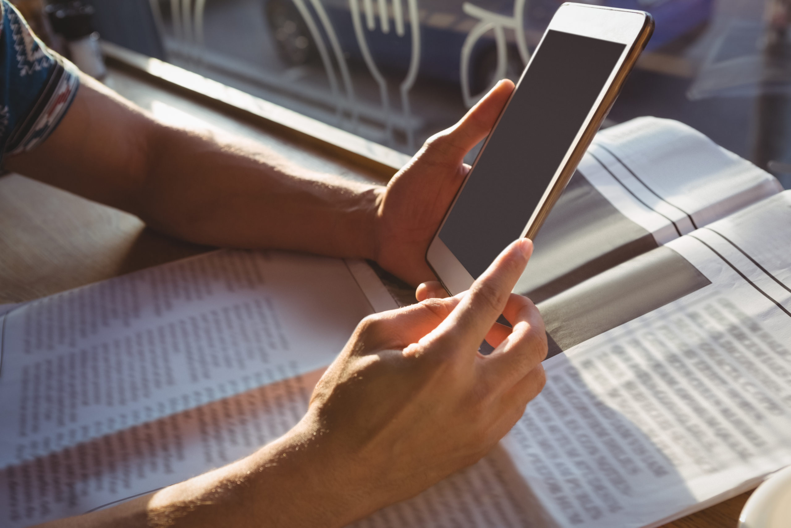 Cropped hands of man with newspaper holding digital tablet by window in cafe